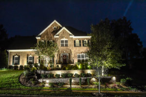 view of large brick two story home at night landscape lighting retaining walls and large trees