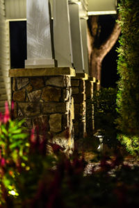 view of front porch columns of home with plants and greenery around stone bases and lighting